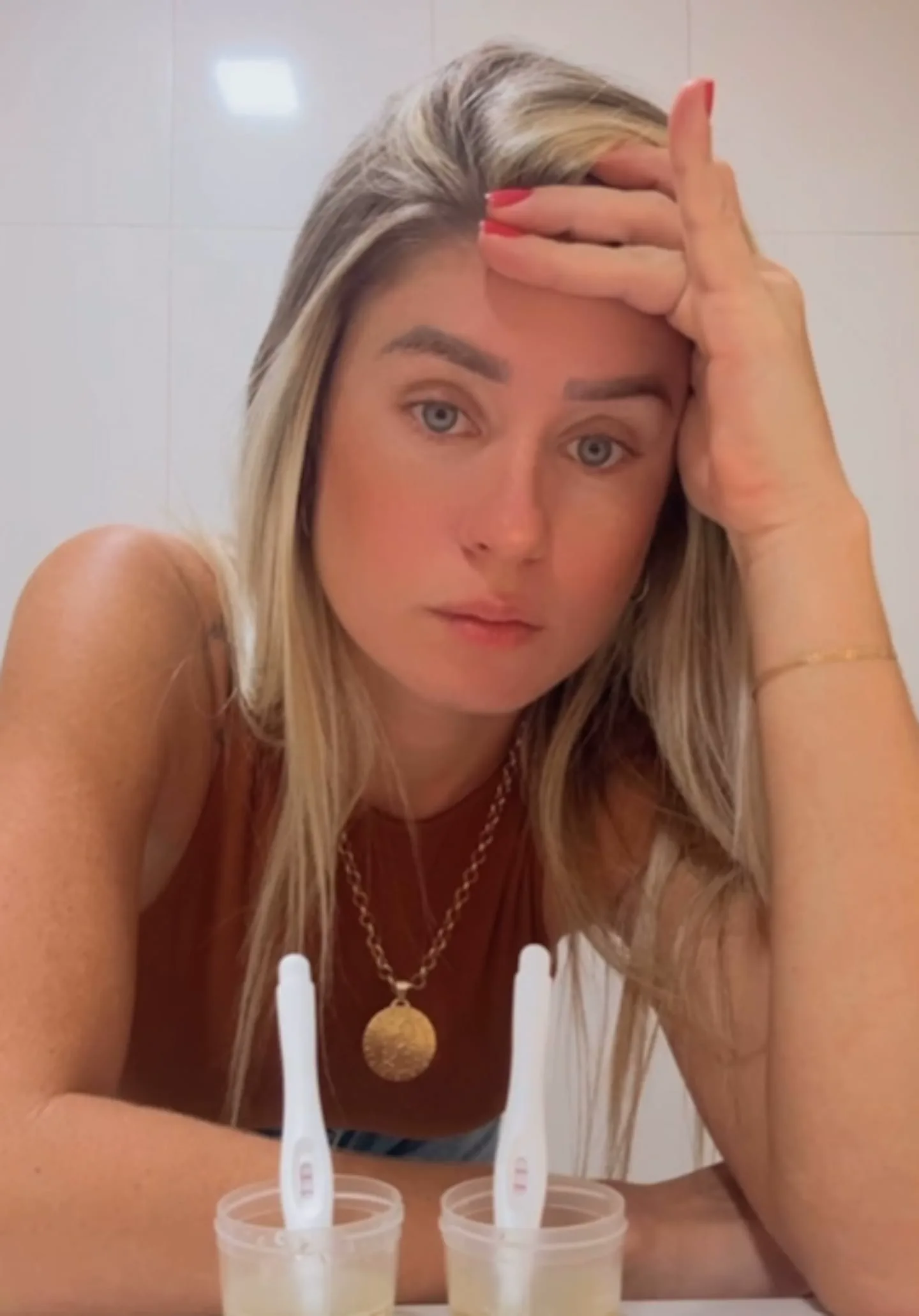 Una mujer rubia apoya la cabeza en su mano frente a dos recipientes con pruebas de embarazo.