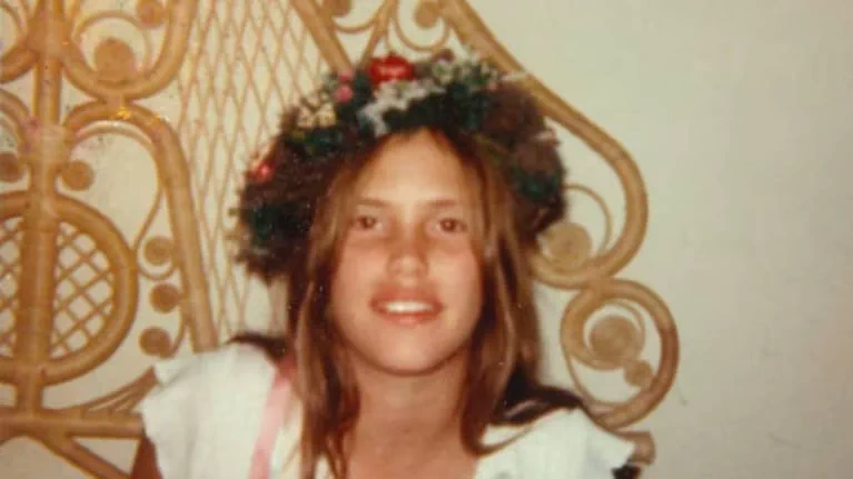 Una chica sonríe con una corona de flores en la cabeza delante de un respaldo de mimbre.