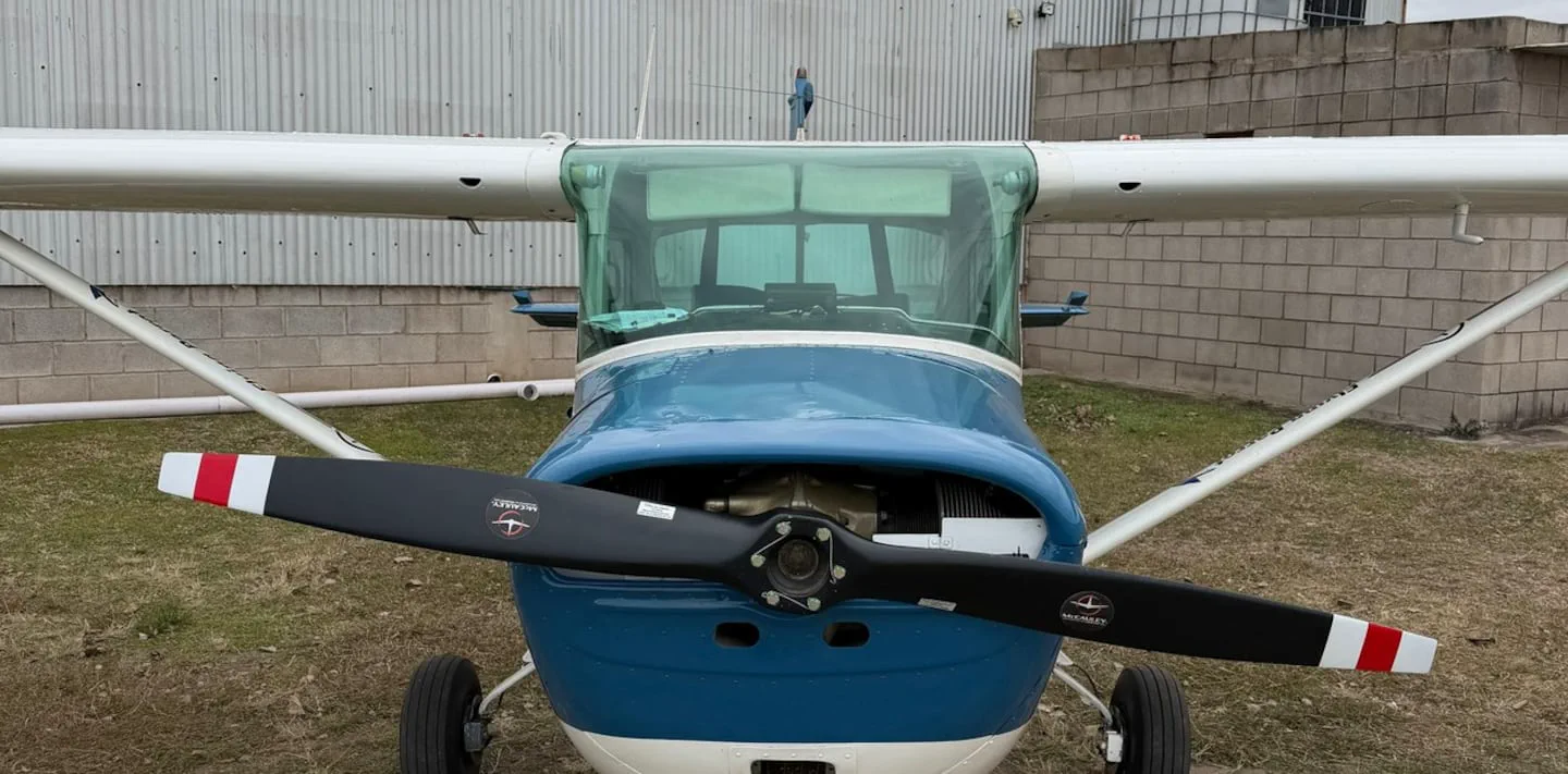 Una avioneta celeste y blanca estacionada de frente en un terreno con pasto seco y un galpon detras.