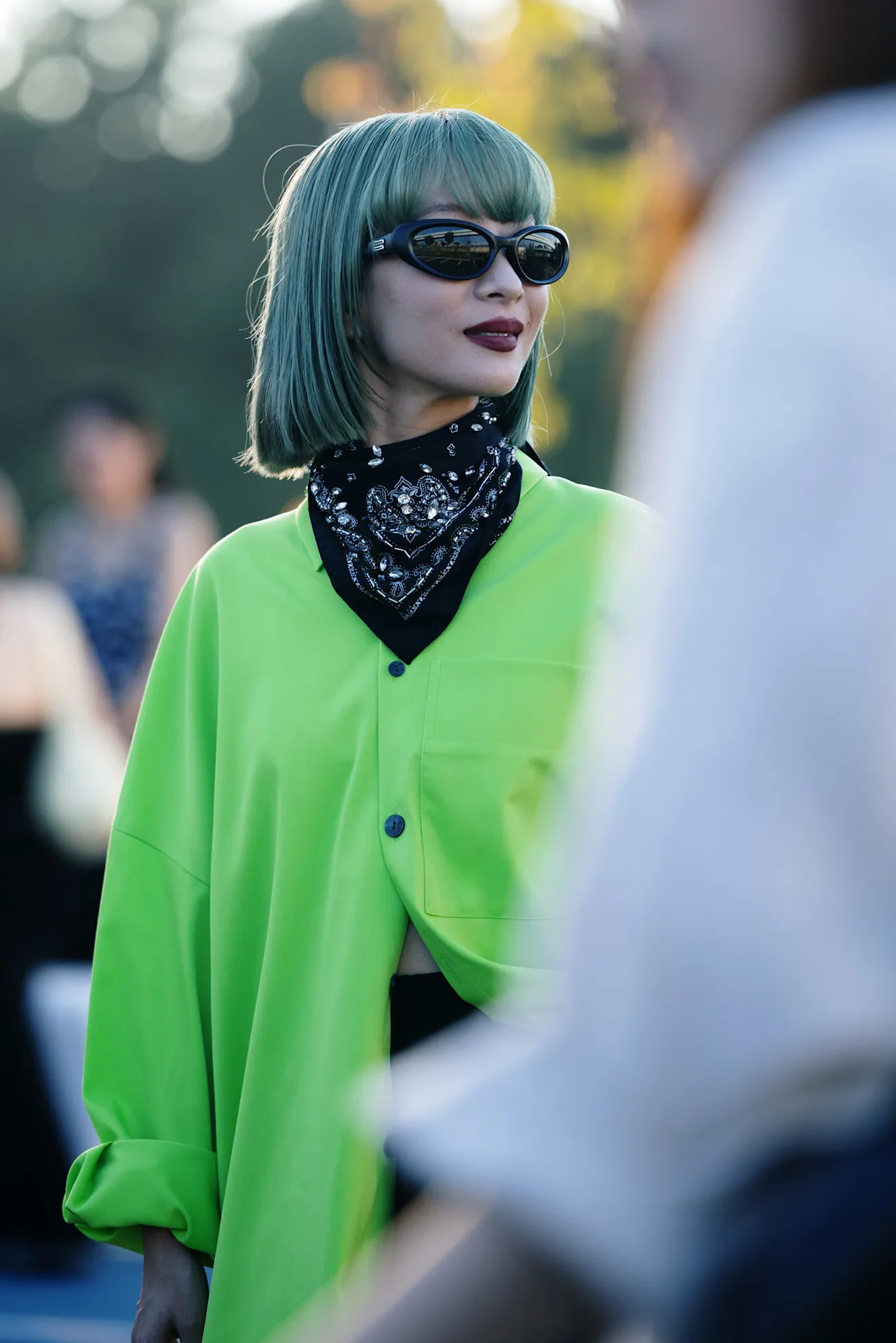 Una persona con pelo teñido verde, anteojos oscuros, camisa verde claro y bandana al cuello.