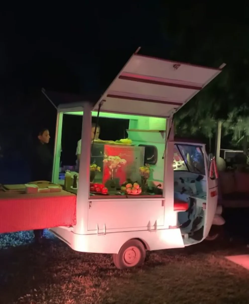 Un motocarro blanco adaptado con barra, frutas, flores y personas atendiendo de noche al aire libre.