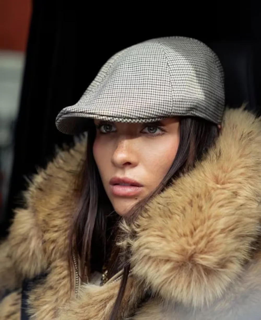 Una mujer de pelo oscuro con boina a cuadros y campera con capucha de piel mirando a cámara.