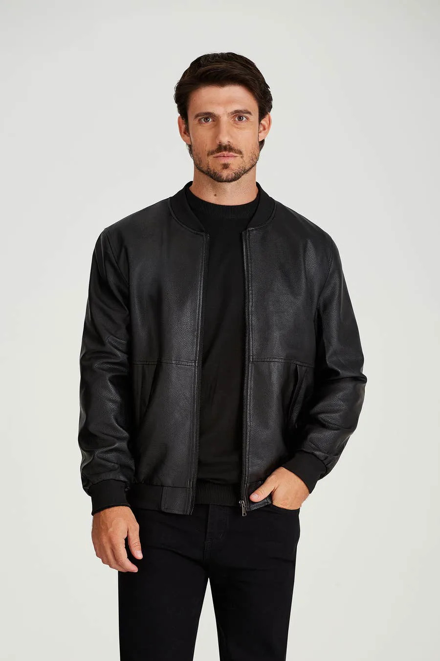 Un hombre barbudo posa de pie vistiendo campera bomber de cuero negro, remera al tono y pantalon oscuro sobre fondo blanco.