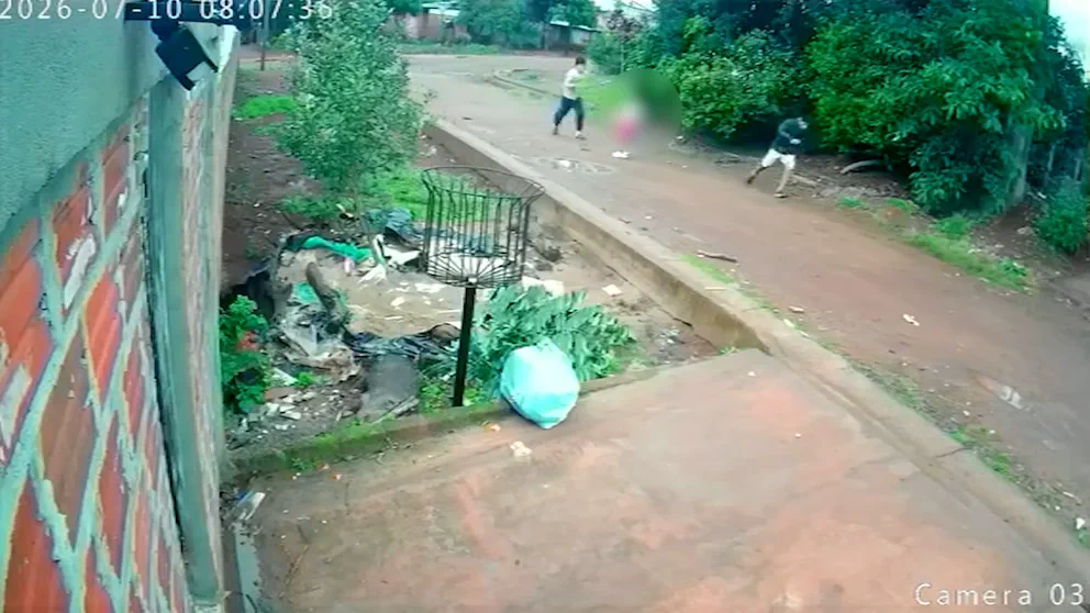 Captura de cámara de seguridad que muestra una calle de tierra con personas a lo lejos y un cesto de basura en primer plano.