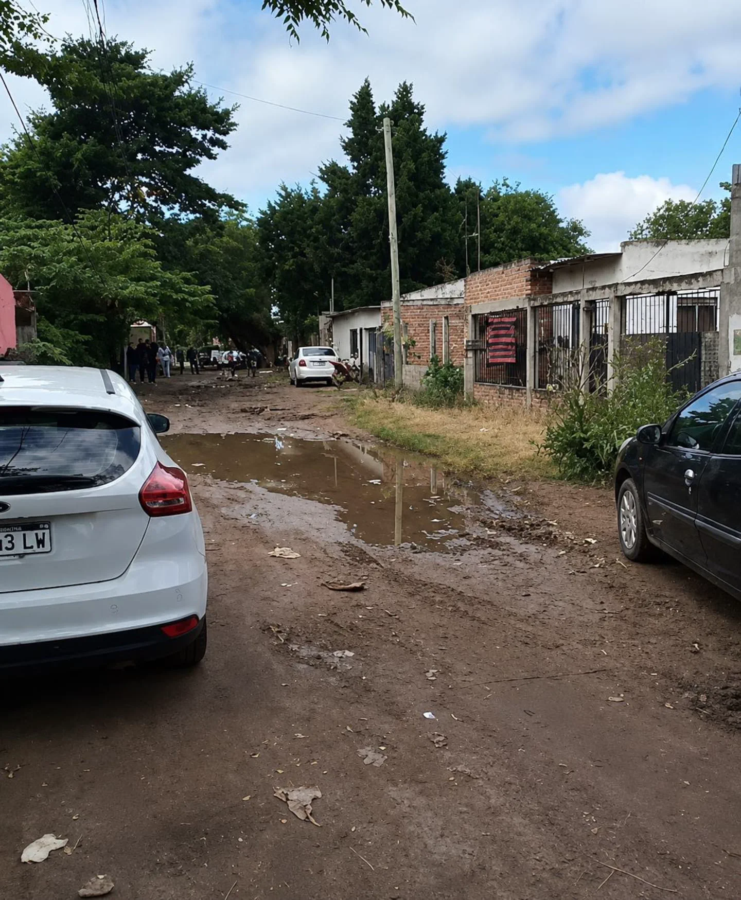 Una calle de tierra con barro y charcos de agua entre casas bajas y autos estacionados.