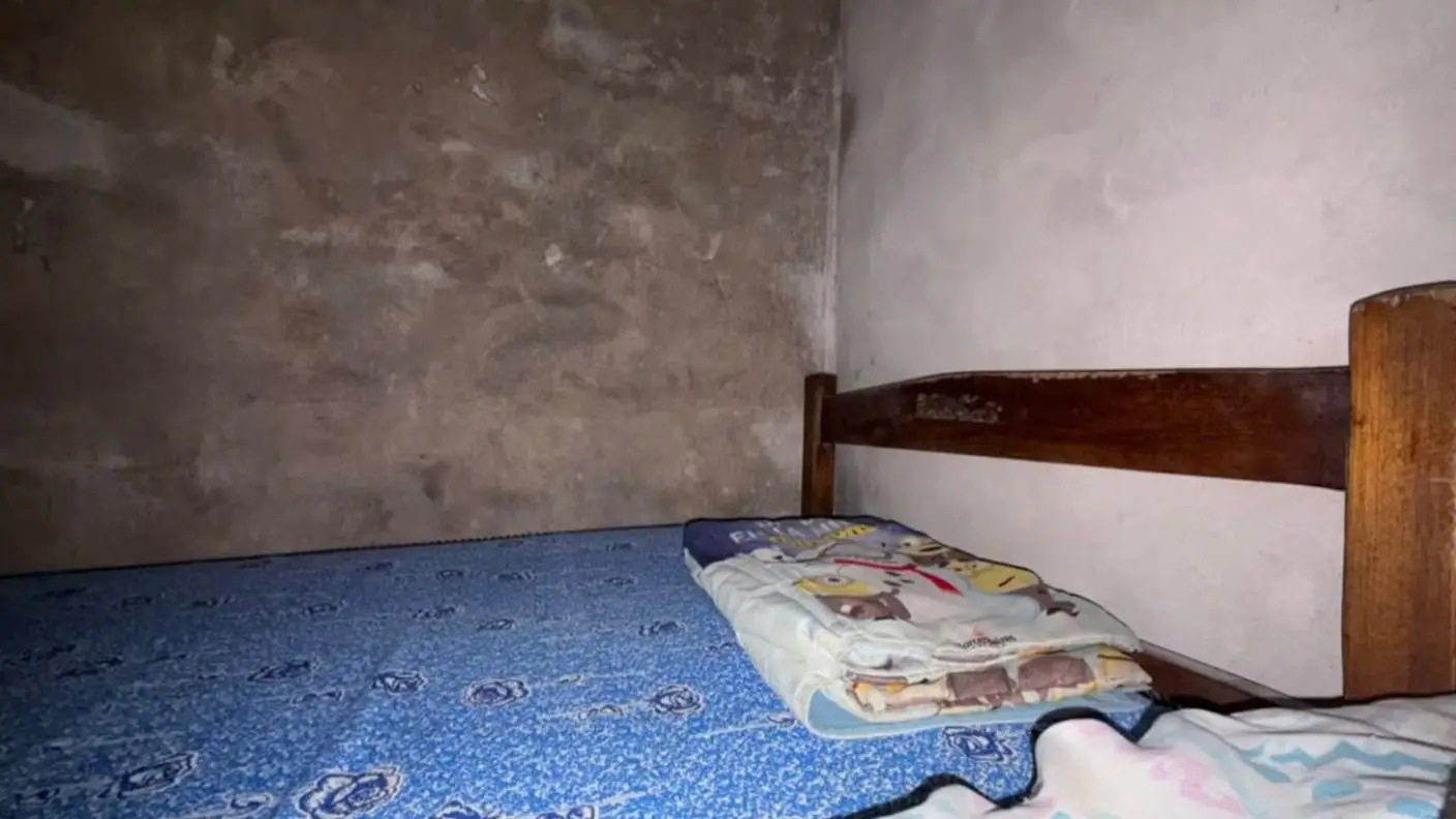 Una cama con colchón azul estampado y frazada doblada está ubicada junto a una pared sin revocar.