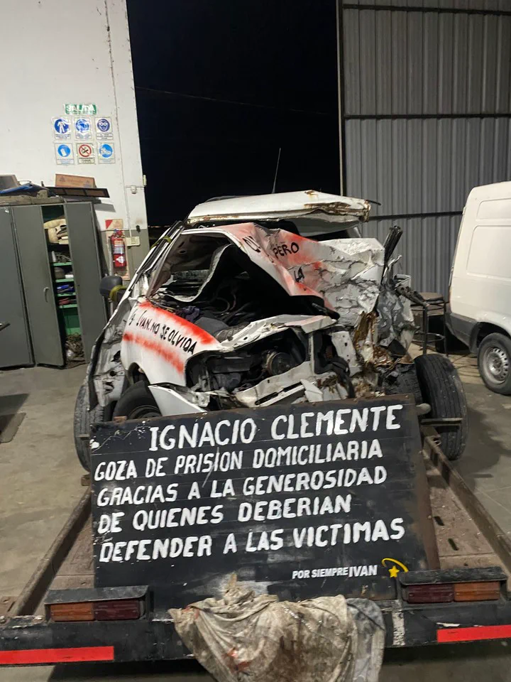Una camioneta completamente destruida por un choque sobre un remolque frente a un cartel de reclamo.