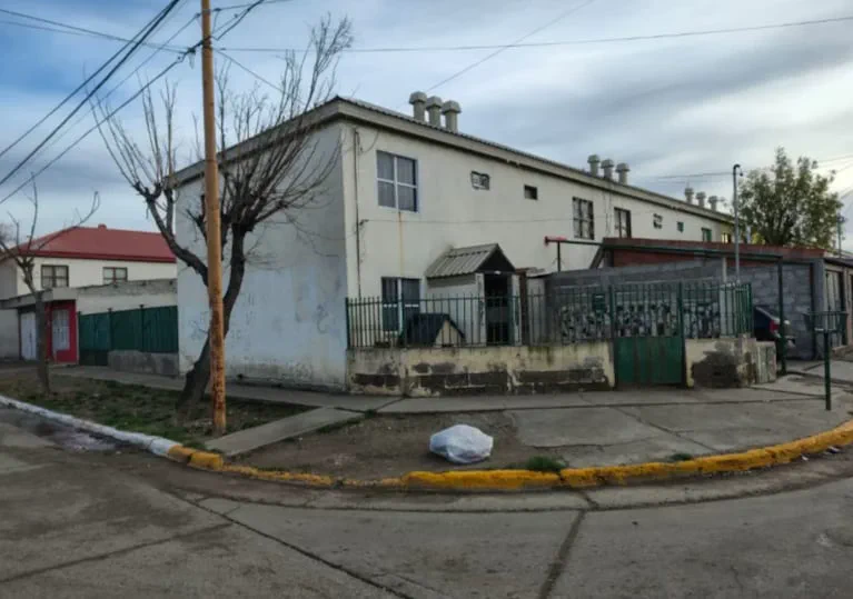 Esquina de un barrio con una casa blanca de dos pisos, rejas verdes y una bolsa de basura en la vereda.