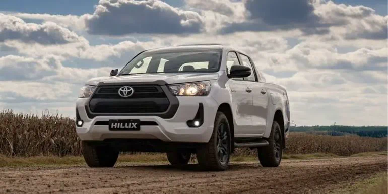 Una camioneta Toyota Hilux blanca esta estacionada sobre un camino de tierra junto a un campo.