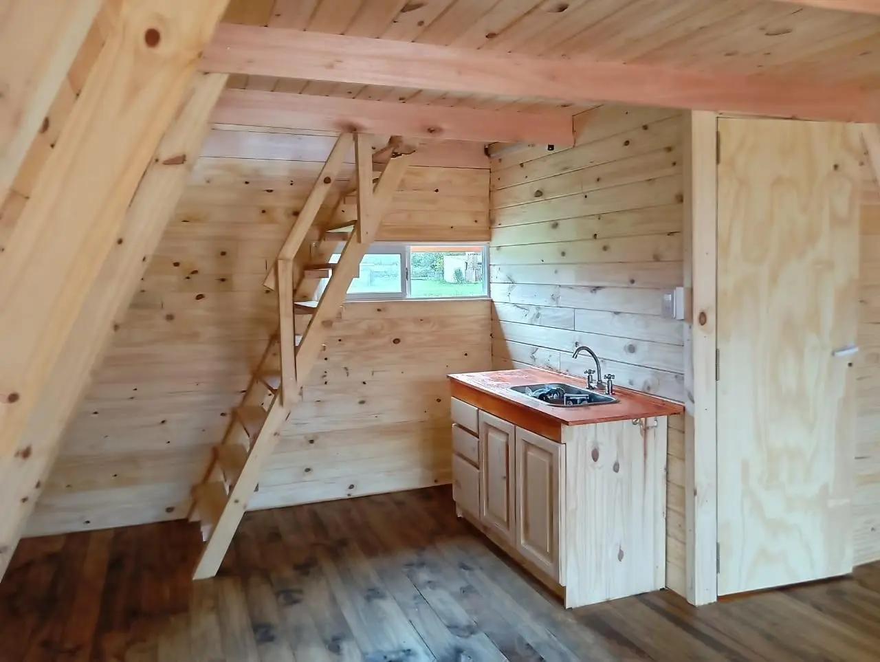 Cocina de madera en una cabaña con mesada, canilla, escalera empinada hacia el altillo y una puerta.
