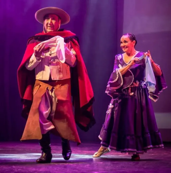 Un hombre con poncho y sombrero y una mujer con vestido violeta bailan folklore sosteniendo pañuelos sobre un escenario.