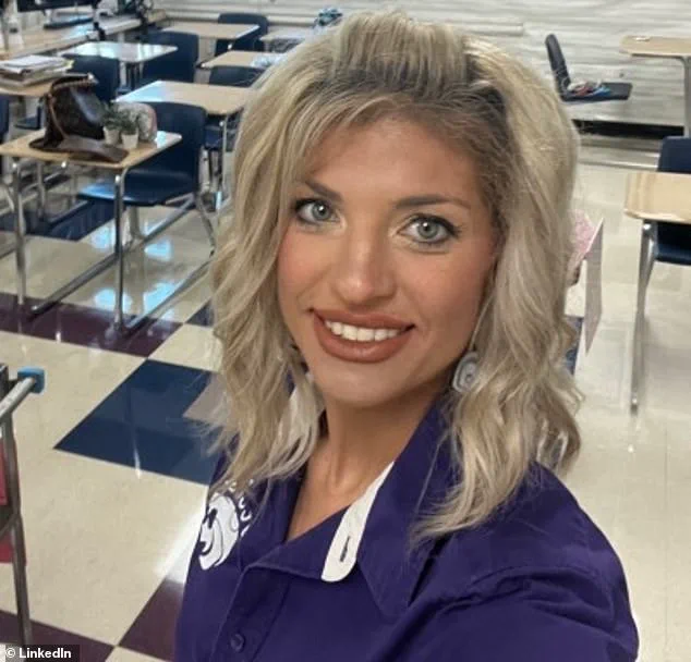 Una mujer rubia y sonriente vestida con chomba violeta se saca una selfie en un aula vacia.