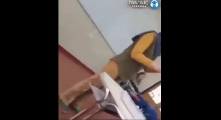 Una persona de espaldas camina por un salón cerca de un escritorio y un pizarrón blanco.