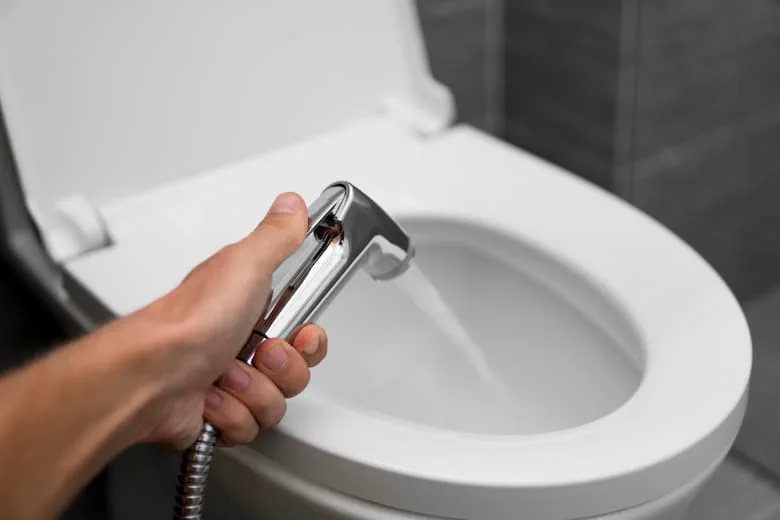 Una mano sostiene una ducha higiénica plateada que tira un chorro de agua hacia el inodoro.