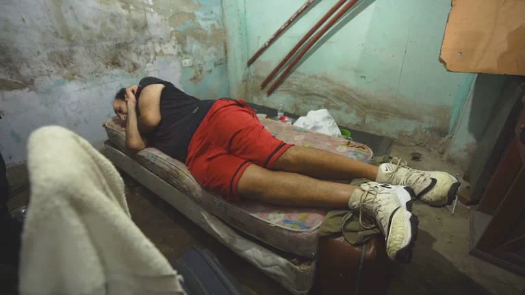 Un hombre recostado boca abajo sobre un colchón en el suelo dentro de una habitación precaria con paredes descascaradas.