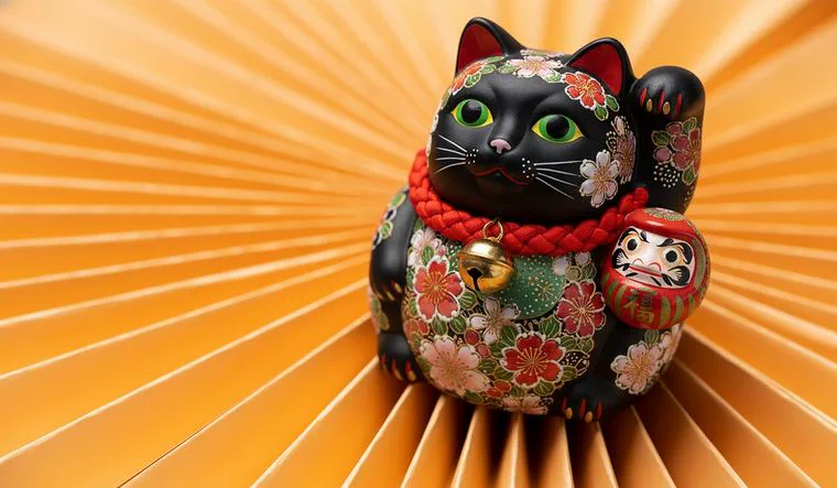Una figura de porcelana de un gato de la suerte negro decorado con flores sobre un abanico plegado color ocre.