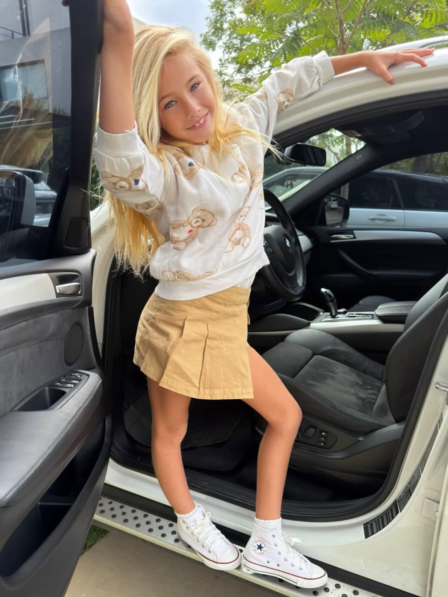 Una nena rubia parada en el estribo de una camioneta blanca con la puerta abierta.