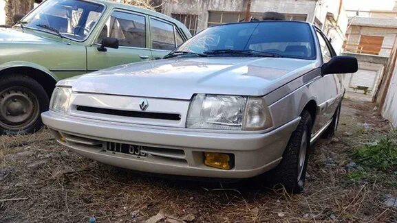 Un auto blanco antiguo estacionado en un patio de tierra junto a otro vehículo.