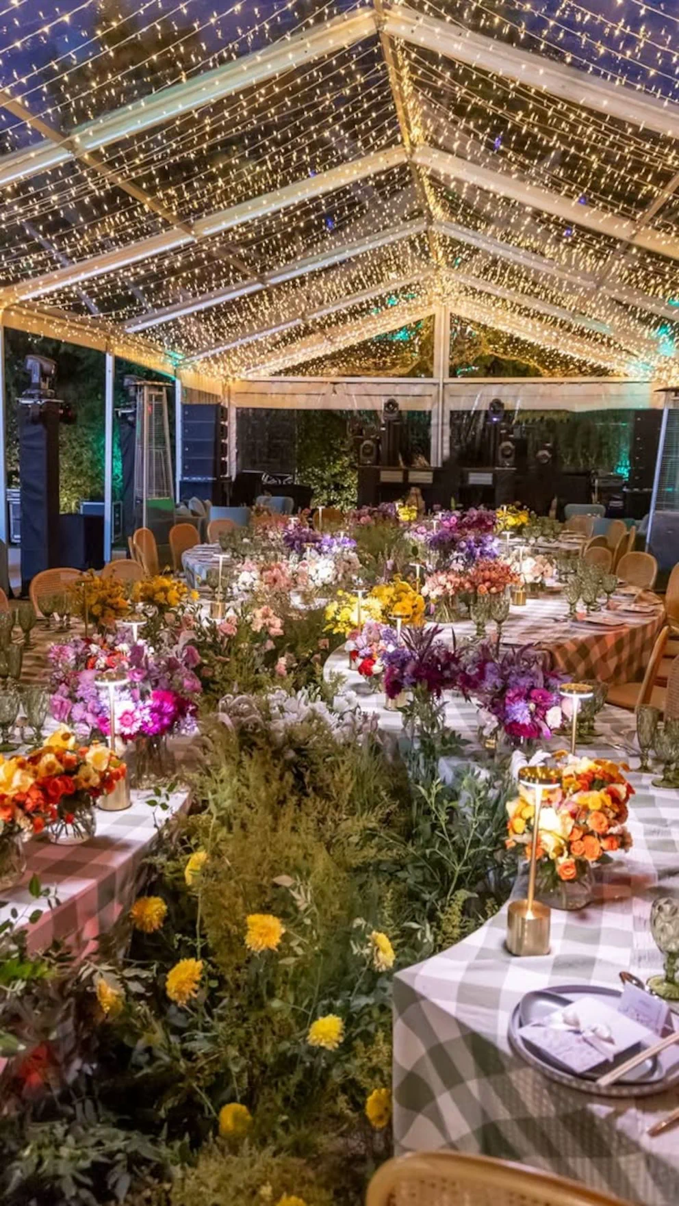 Mesas largas decoradas con flores, vajilla y luces cálidas distribuidas bajo el techo de una carpa.