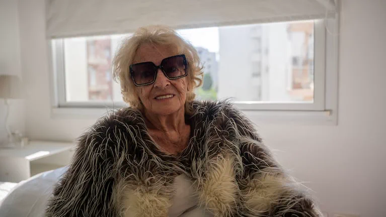 Mujer mayor con tapado de plumas y anteojos de sol oscuros sentada frente a una ventana.