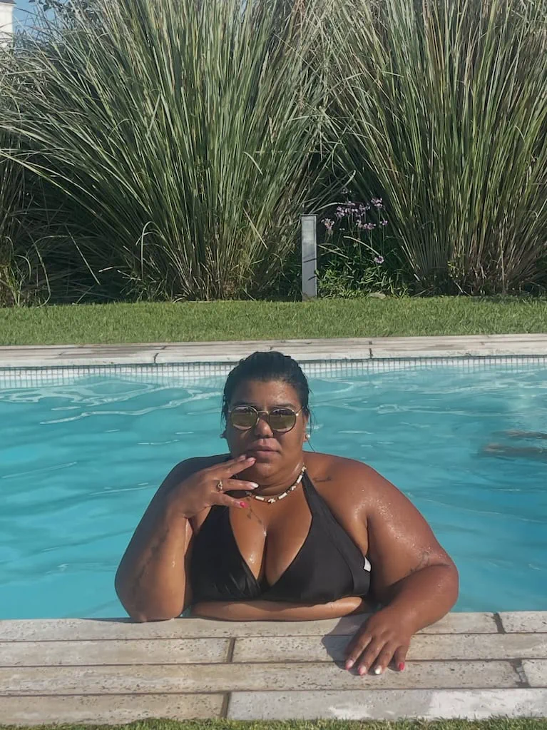 Una mujer con anteojos oscuros y bikini negra posa apoyada sobre el borde de una pileta.