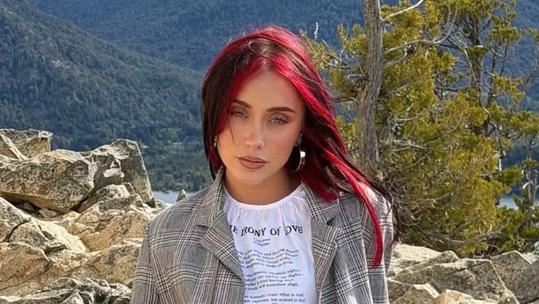 Mujer joven con pelo teñido de rojo y campera a cuadros posando al aire libre frente a un paisaje montañoso.