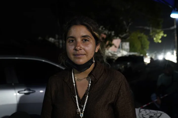 Una mujer joven con el barbijo bajo y collares mira a la camara al aire libre de noche.