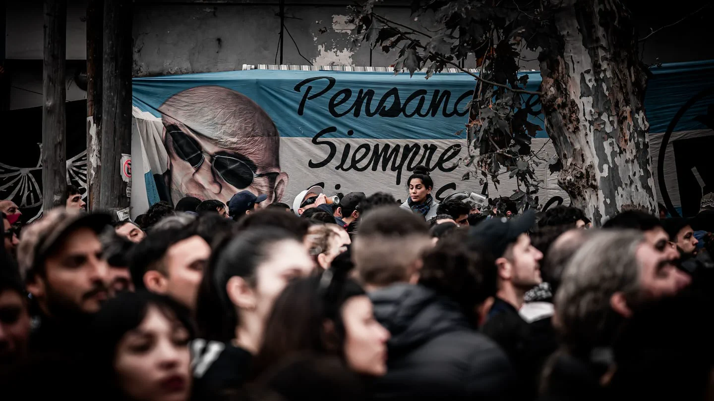 Una multitud reunida al aire libre frente a una bandera que lleva pintado el rostro del Indio Solari con lentes de sol.