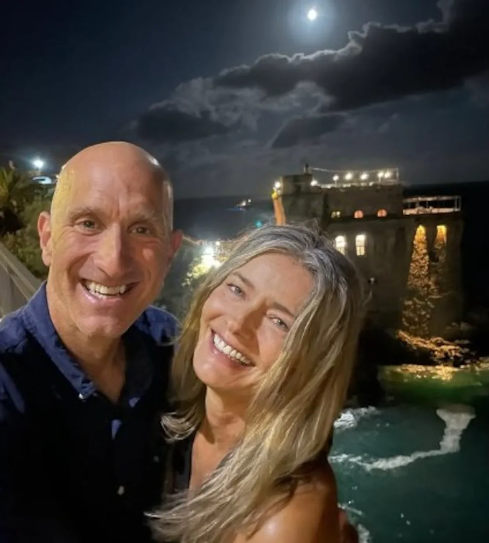 Un hombre calvo y una mujer rubia sonríen de noche con una edificación iluminada y el mar de fondo.