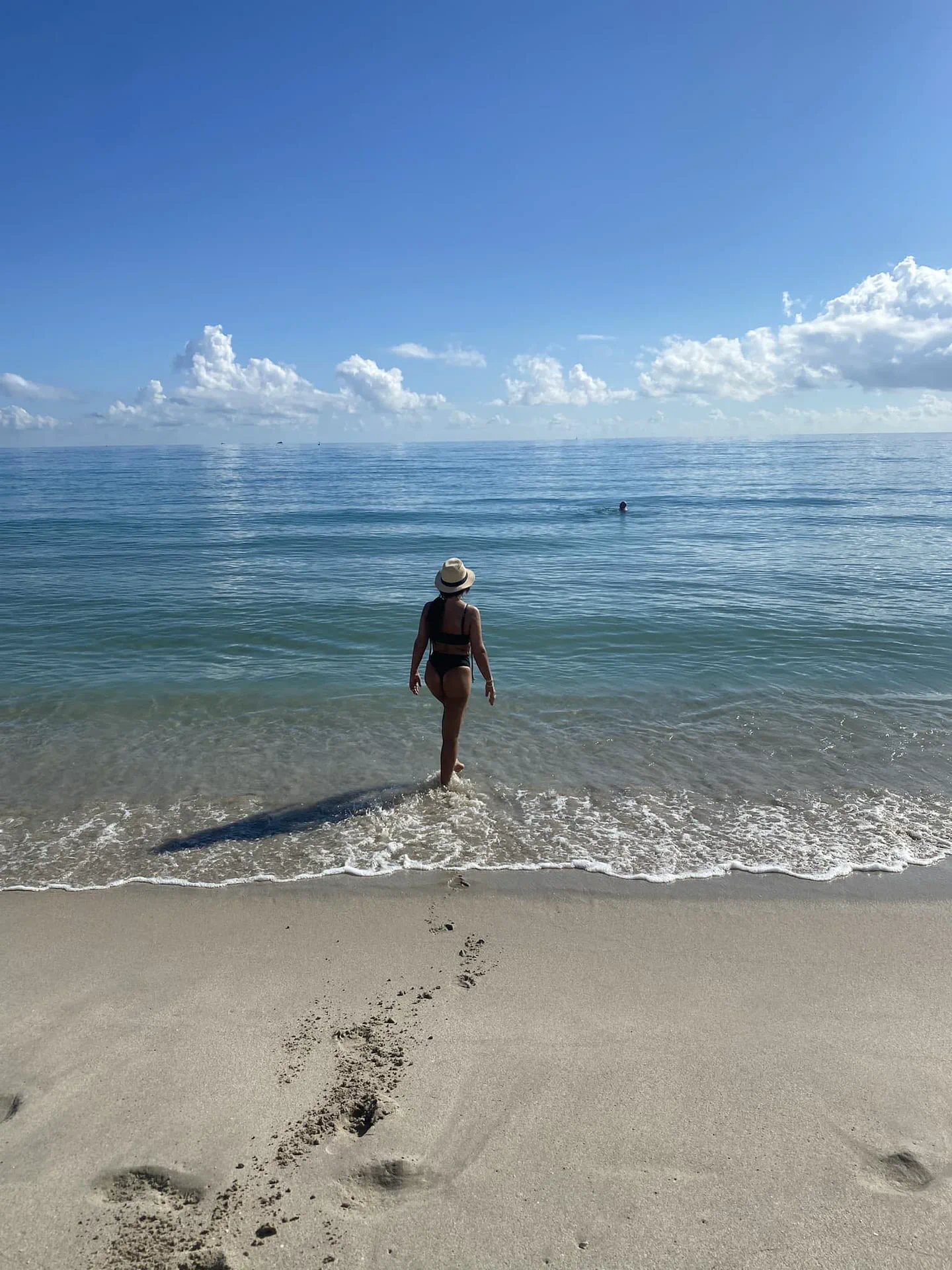 Una mujer en bikini y sombrero camina de espaldas hacia el agua en una playa de mar calmo y arena clara.