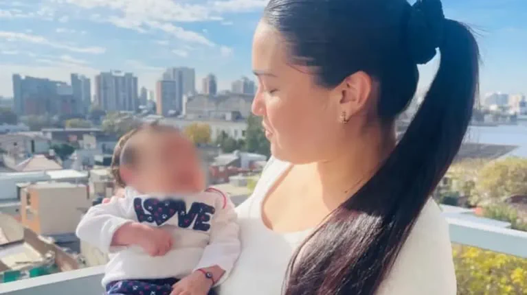 Una mujer con pelo recogido sostiene en brazos a un bebe con la cara pixelada en un balcon con edificios detras.