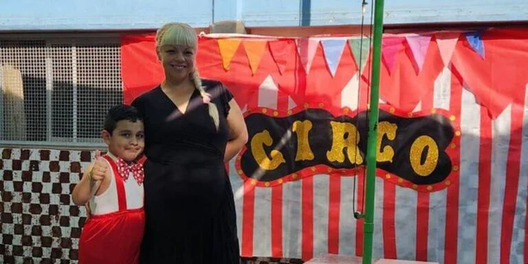 Una mujer rubia y un nene con mono rojo posan sonrientes frente a una escenografia de circo.