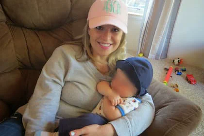 Una mujer con gorra rosa sostiene en brazos a un bebé con gorro azul en un sillón dentro de una casa.
