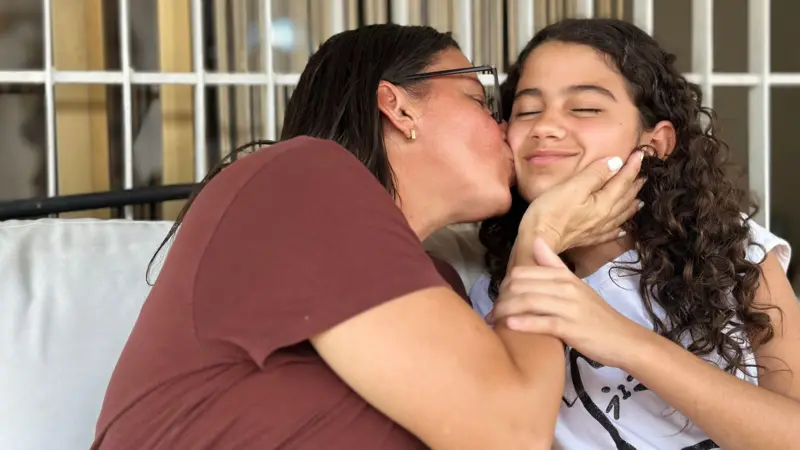 Una mujer con anteojos le da un beso en la mejilla a una chica de pelo enrulado que sonríe.