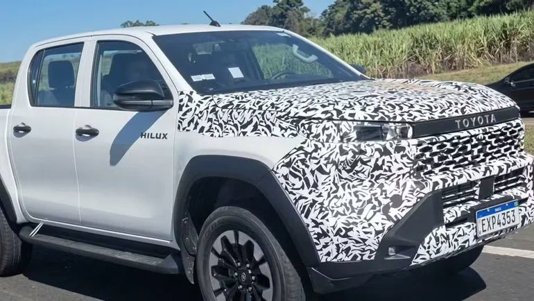 Una camioneta Toyota Hilux blanca con ploteo de camuflaje en la trompa circula por una ruta.