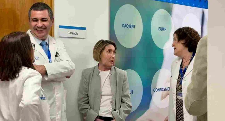 Una mujer en saco claro conversa en un pasillo con varios médicos con delantal blanco.