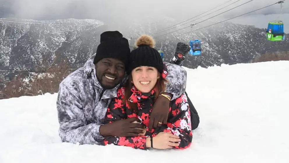Una pareja recostada en la nieve posa sonriendo con un cerro nevado y cabinas de teleférico de fondo.