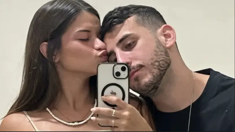 Una chica le da un beso en la mejilla a un muchacho mientras se sacan una selfie frente al espejo con un celular.