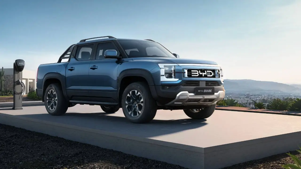 Una camioneta pickup azul estacionada en una plataforma exterior con un cargador eléctrico al lado.