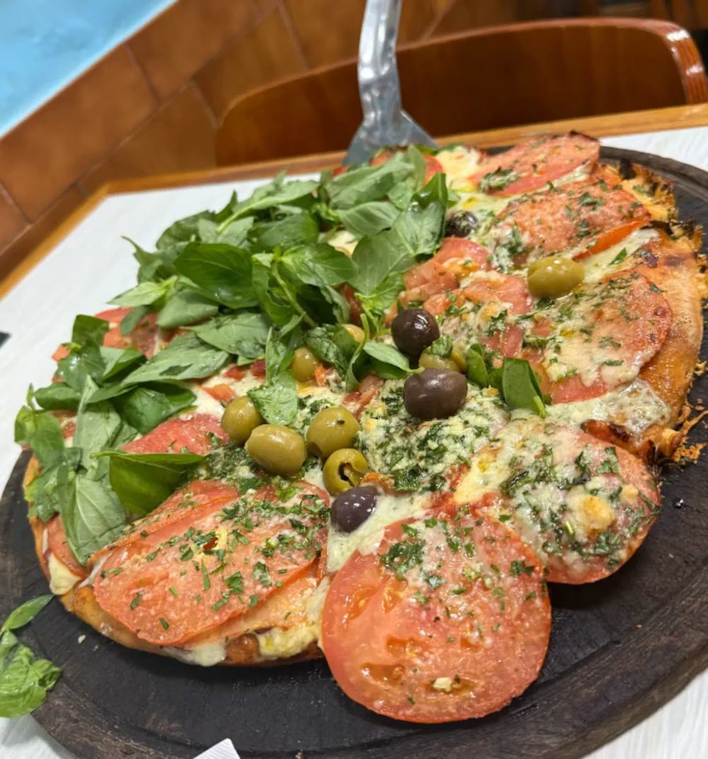 Una pizza servida sobre una tabla de madera redonda con rodajas de tomate, aceitunas y abundantes hojas de albahaca.
