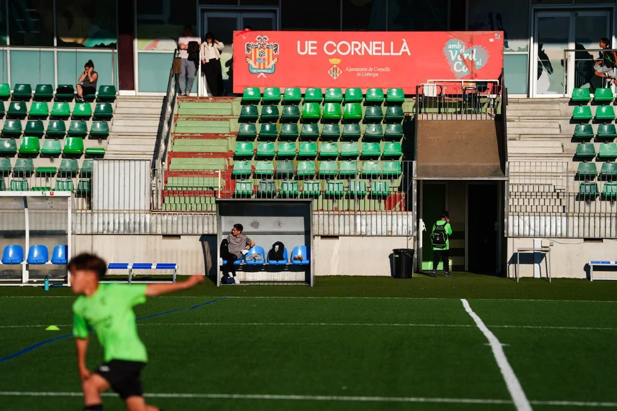 Tribuna del club UE Cornella con butacas verdes y rojas y personas en la platea y el banco de suplentes.