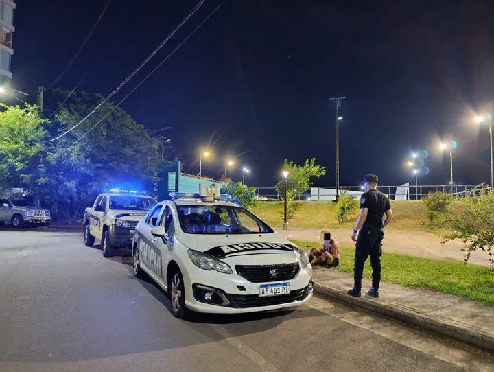 Un policía de espaldas junto a dos patrulleros y un hombre sentado en el cordón de noche.