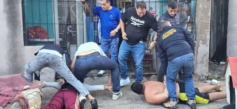 Varios efectivos policiales reducen a dos hombres acostados boca abajo sobre la vereda frente a una reja.