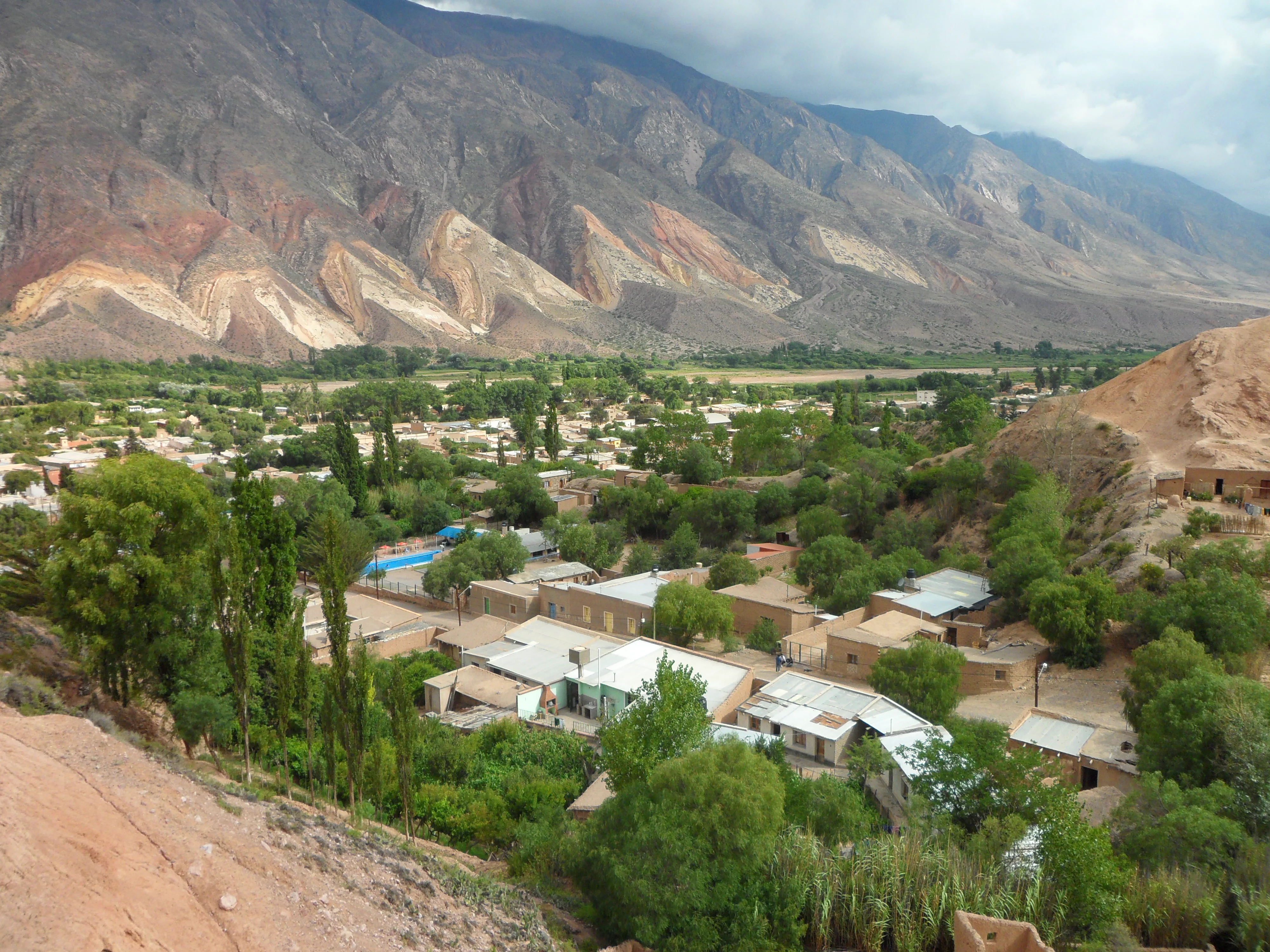 Vista panorámica de un pueblo entre árboles con cerros de colores de fondo.