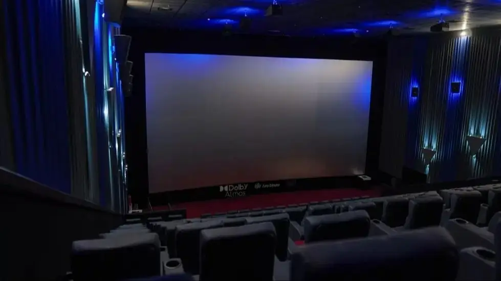 Una sala de cine vacía con butacas oscuras y una gran pantalla proyectando un fondo gris iluminado.