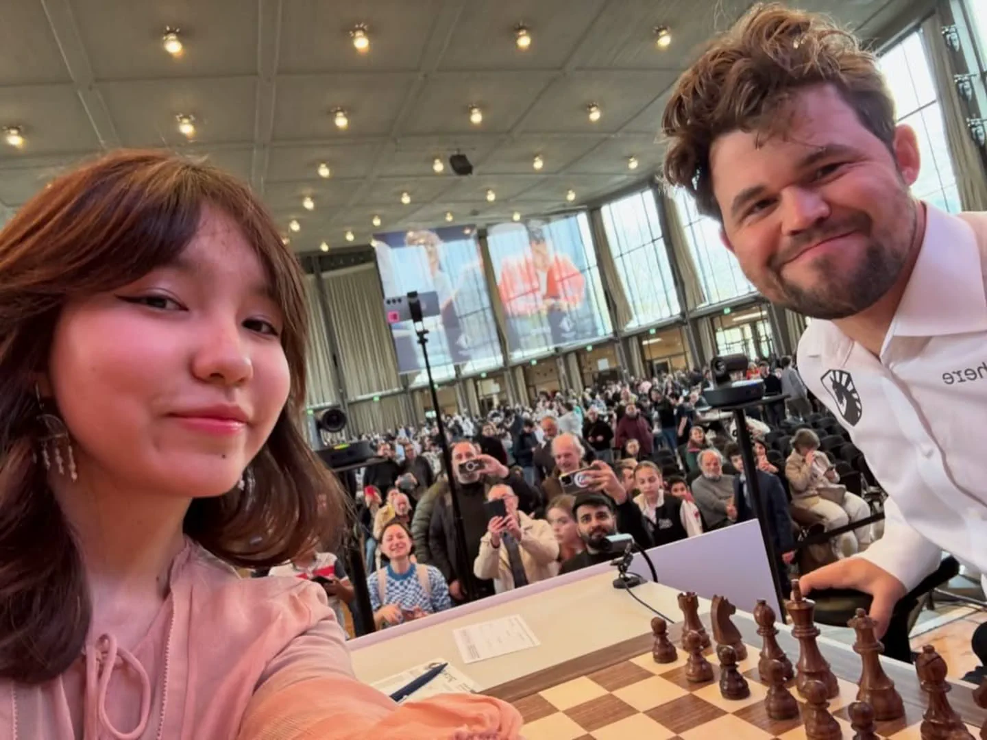 Una joven y el ajedrecista Magnus Carlsen posan para una selfie junto a un tablero con público detrás.