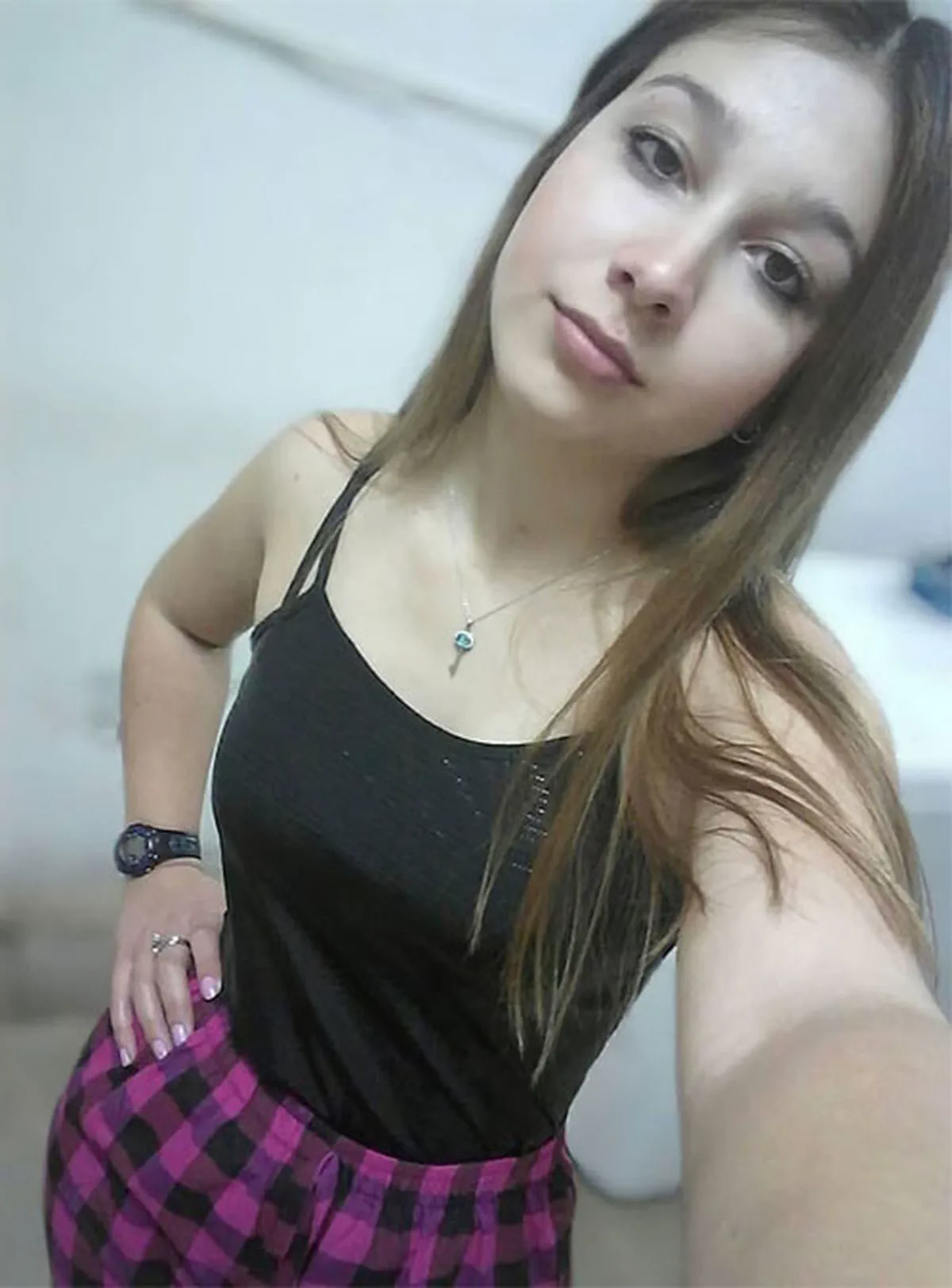 Una chica joven con musculosa negra y pantalón cuadrillé se saca una selfie inclinando la cabeza.