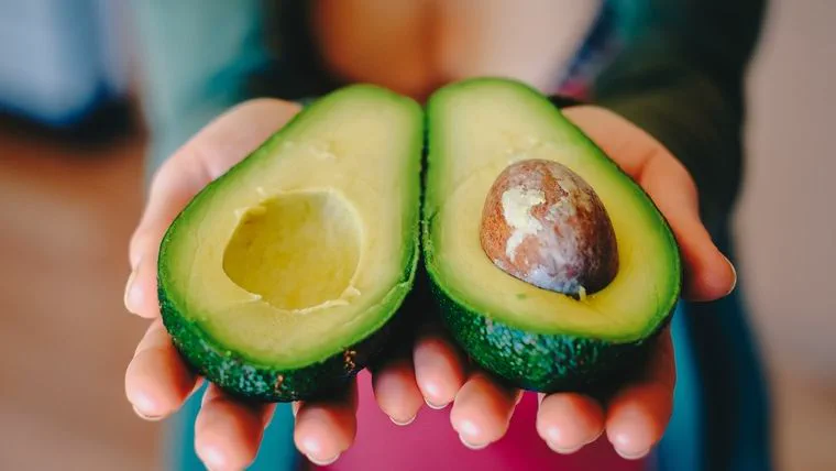 Dos manos sostienen dos mitades de una palta abierta que deja ver el carozo.