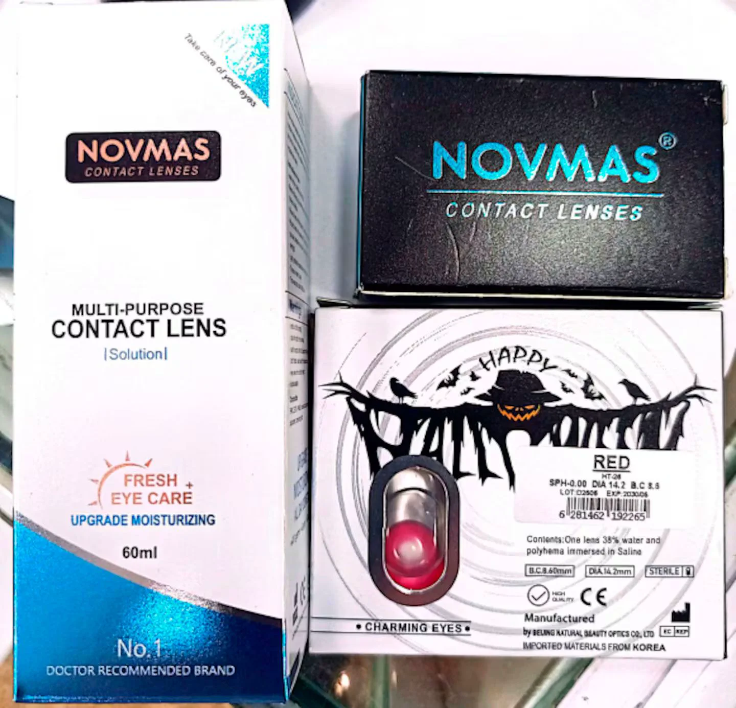 Caja de solución para lentes de contacto Novmas junto a un blister con una lente de contacto de color rojo.