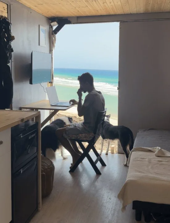 Persona sentada frente a una notebook junto a dos perros mirando hacia la playa desde el interior de una casilla.